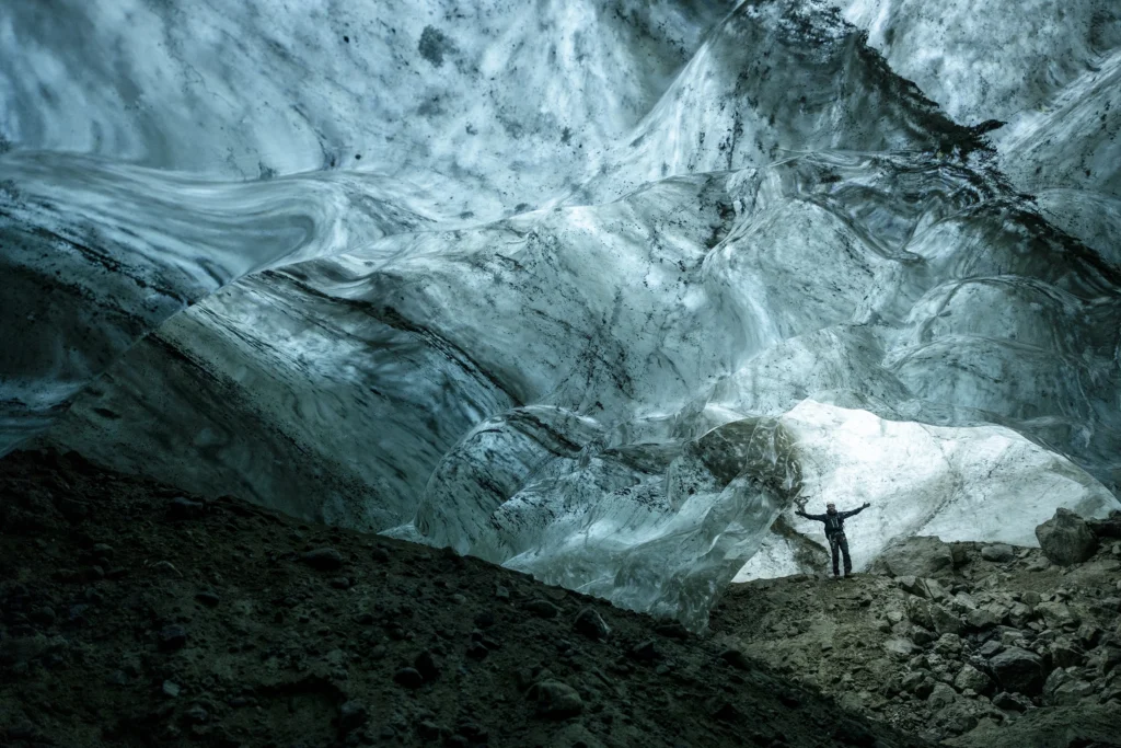 Person standing inside a large ice cave in East Greenland, surrounded by smooth blue ice formations. Photo by Oscar Scott Carl - Visit East Greenland