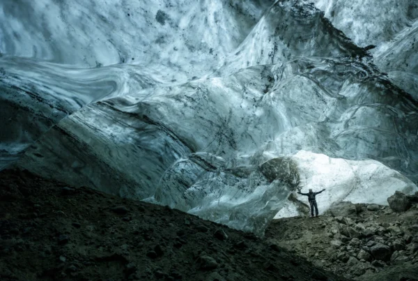 Person standing inside a large ice cave in East Greenland, surrounded by smooth blue ice formations.