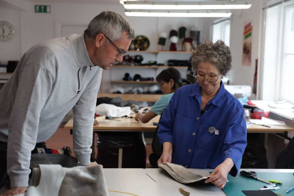 A woman showing her work progress to a curious tourist at the local sealskin sewing facility.