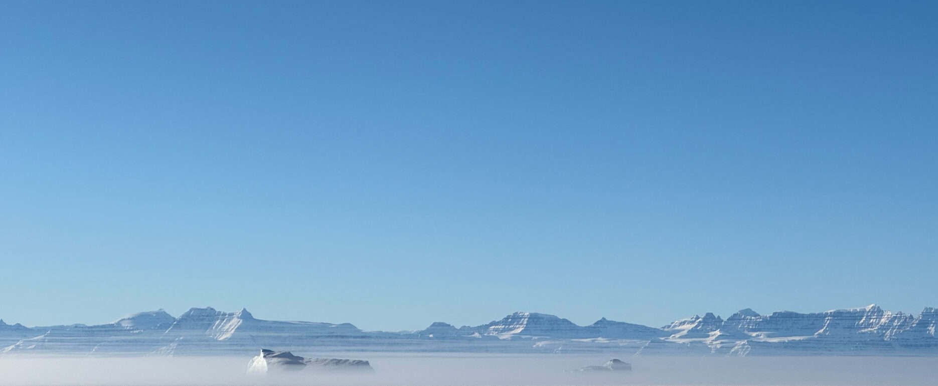 Fog rolling in over the sea ice packed fjord and the distant mountains of the Scoresbysund area.